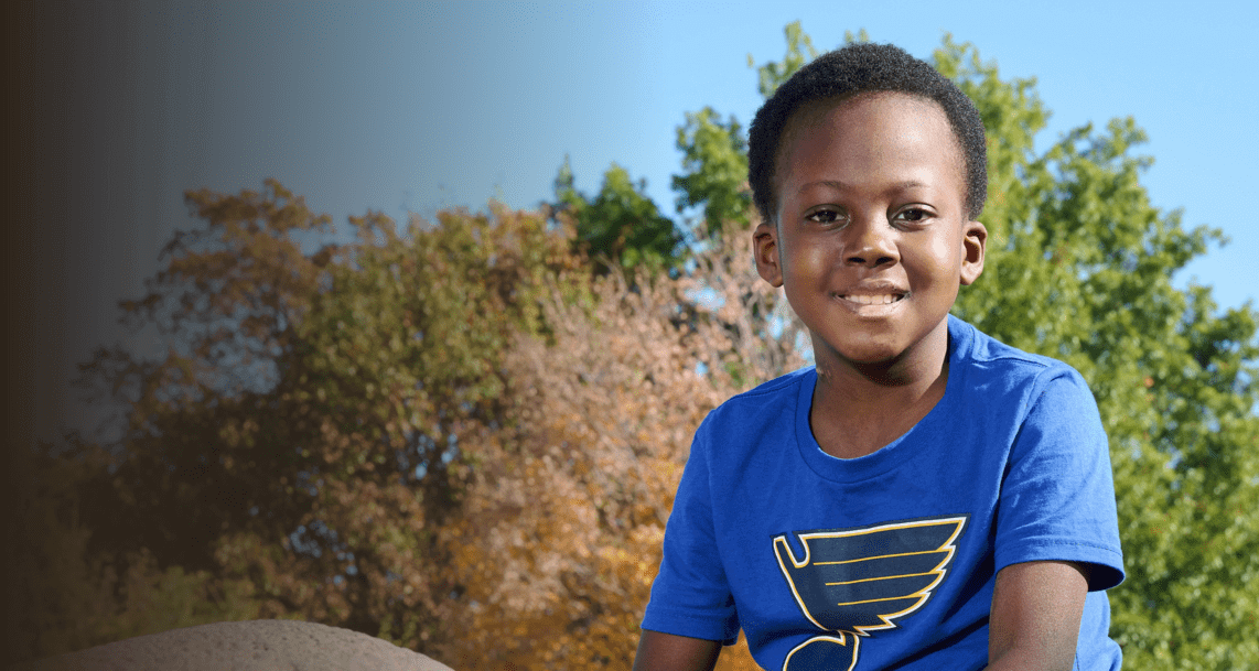 A 7-year-old boy in a St Louis Blues shirt sits on a statue at a park. The trees behind him are starting to turn to fall colors.