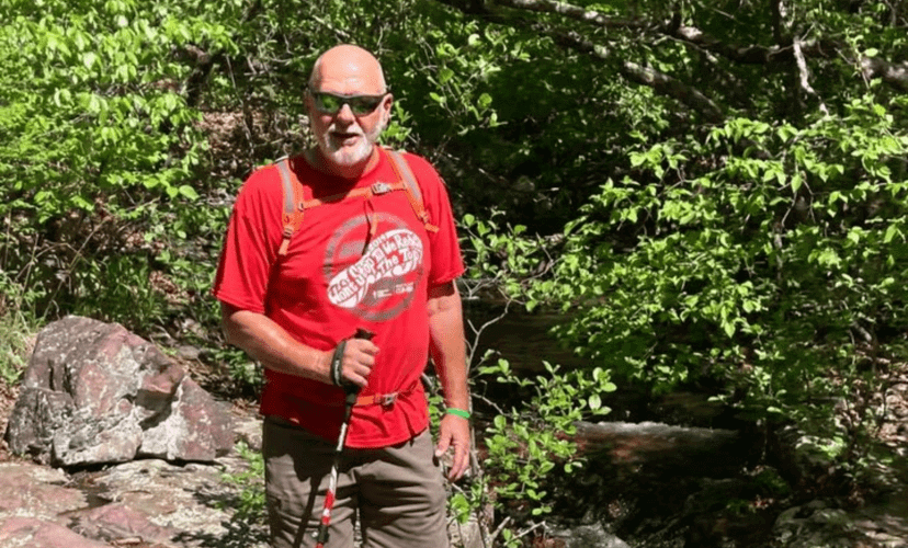 A man is hiking in the woods. He is wearing a sunglasses, a red shirt, and a green Donate Life bracelet. He is holding a hiking stick.