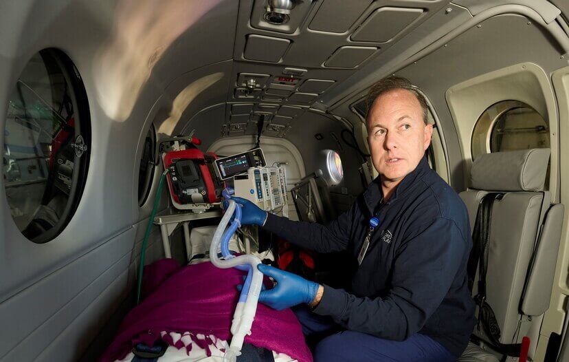 A man sitting inside an airplane wears blue latex gloves and holds a ventilator tube. The patient in front of him is covered in a purple blanket.