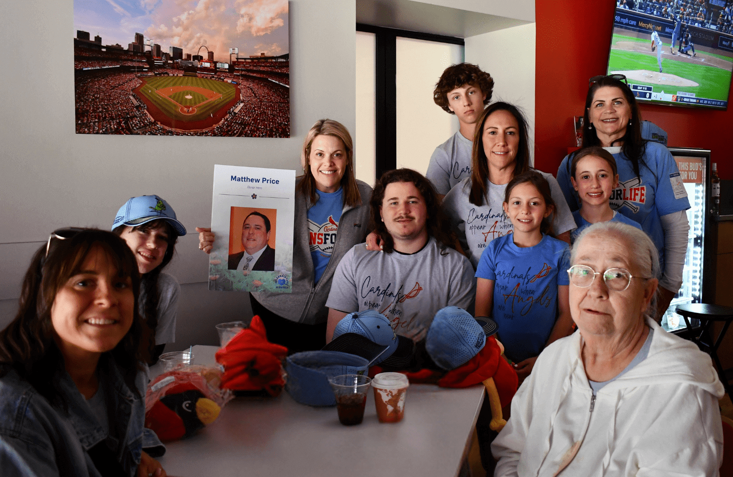 The Price family gathers around a table at Busch Stadium, along with two Mid-America Transplant Employees. One employee holds up a sign with a photo of Matthew Price on it. Matthew became a tissue donor in 2023.