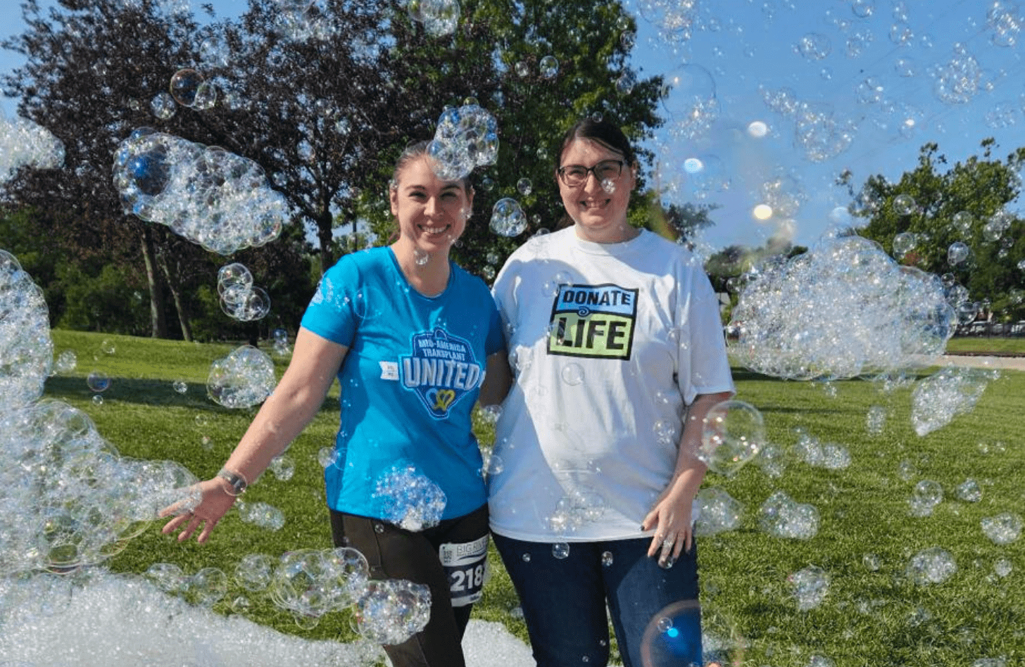 Two women stand in a field surrounded by bubbles. Deb is wearing a shirt that says "Mid-America Transplant United". Her sister Melissa wears a white Donate Life shirt.