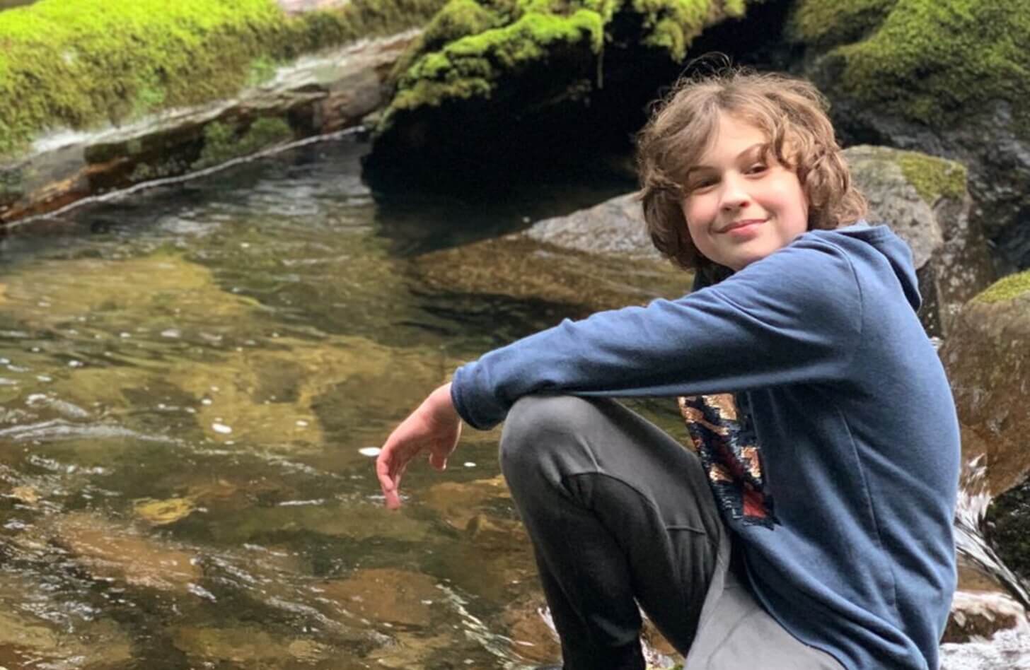 Beckham, a teenager with brown hair and a blue hoodie, sits on a rock next to a creek